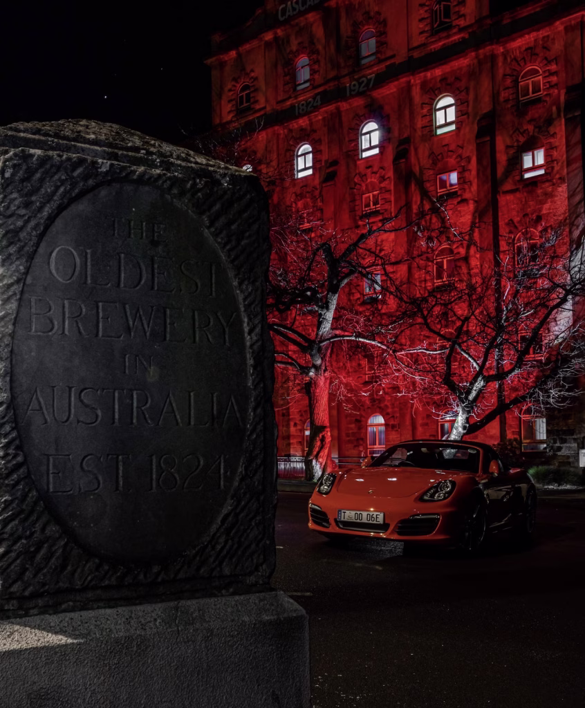 A red Porsche from Drive Car Hire stands parked next to a gravestone reading "The Oldest Brewery in Australia est. 1821" in a moody shot. The building behind is lit up with red lights for the Dark Mofo winter festival.